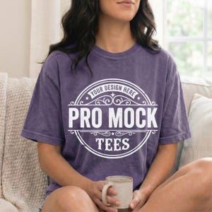May include: A purple t-shirt with the text "YOUR DESIGN HERE PRO MOCK TEES" in a white circular design. The person is holding a beige mug. The shirt has short sleeves and a crew neck.