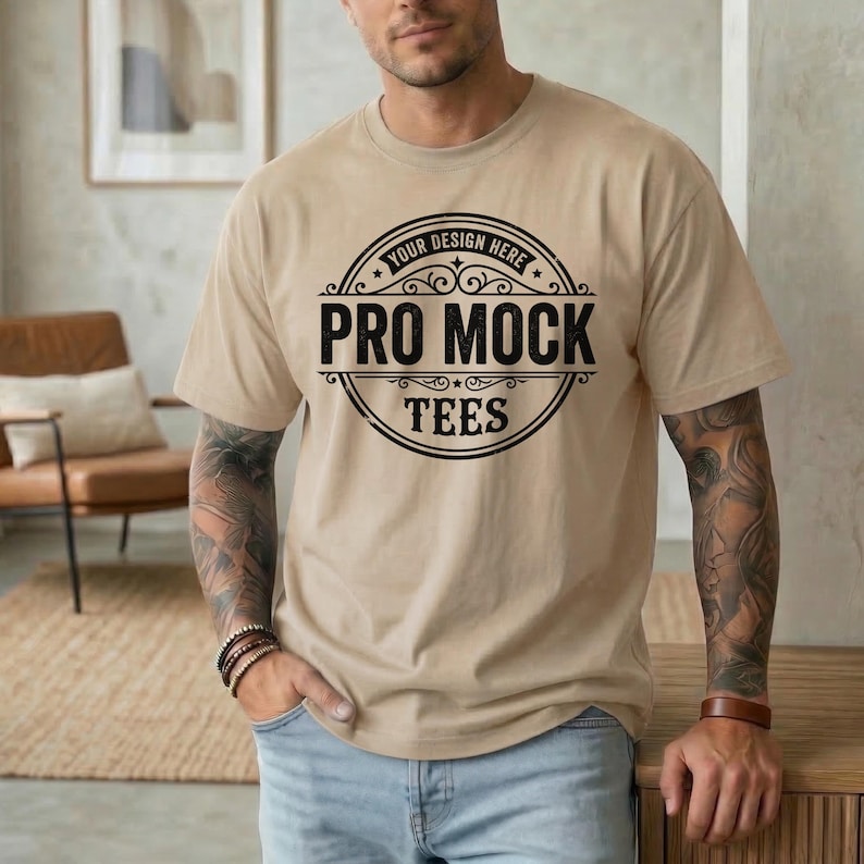 May include: A tan t-shirt with the text "PRO MOCK TEES" in a black circular design. The shirt is being worn by a person in blue jeans. The text "YOUR DESIGN HERE" is above the main text. The person has tattoos on their arms.
