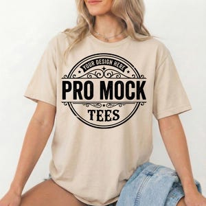 Peut inclure: T-shirt beige avec un motif circulaire noir. Le motif comprend le texte "YOUR DESIGN HERE" en haut et "PRO MOCK TEES" en lettres noires en gras. Le t-shirt a des manches courtes et un col rond.