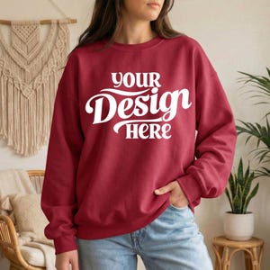 May include: A maroon sweatshirt with the text "YOUR Design HERE" in white script. The sweatshirt is a solid color and has long sleeves. The model is wearing blue jeans.