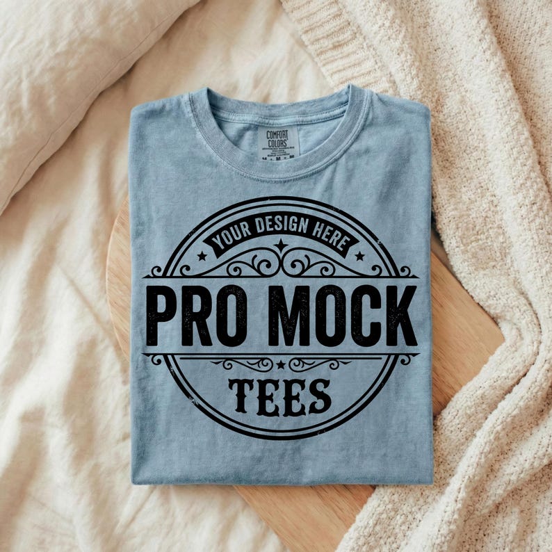Puede incluir: Camiseta azul claro con cuello redondo, doblada cuidadosamente. Presenta un dise&ntilde;o circular negro con el texto "YOUR DESIGN HERE" y "PRO MOCK TEES". La camiseta est&aacute; sobre una superficie de madera, con una manta crema de fondo.