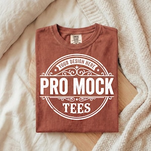 Puede incluir: Una camiseta doblada de color &oacute;xido con un gr&aacute;fico circular blanco que dice "YOUR DESIGN HERE PRO MOCK TEES". La camiseta est&aacute; hecha de un material suave y tiene cuello redondo.