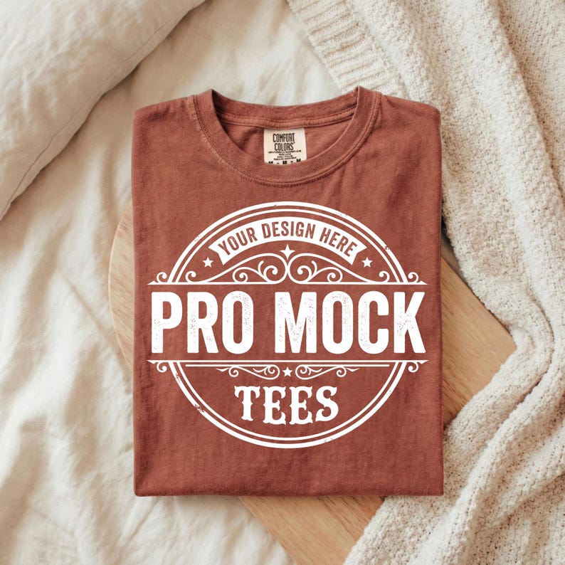 Puede incluir: Una camiseta doblada de color &oacute;xido con un gr&aacute;fico circular blanco que dice "YOUR DESIGN HERE PRO MOCK TEES". La camiseta est&aacute; hecha de un material suave y tiene cuello redondo.