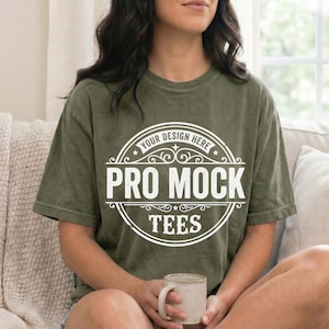 May include: Olive green t-shirt with the text "YOUR DESIGN HERE" and "PRO MOCK TEES" in a white circular design. The model is holding a beige mug. The shirt is a short-sleeved, casual style.