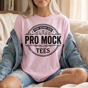 May include: A light pink t-shirt with a black circular design that reads "Your Design Here" at the top, "Pro Mock" in the center, and "Tees" at the bottom. The shirt is worn with a denim jacket and blue jeans.
