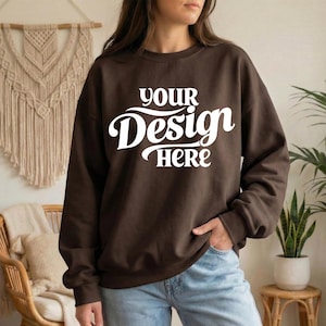 May include: A brown sweatshirt with the text "YOUR Design HERE" in white script. The sweatshirt is a solid color and has long sleeves. The model is wearing blue jeans.