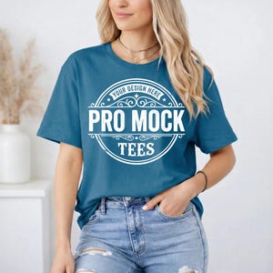 May include: Teal-colored t-shirt with the text "PRO MOCK TEES" in a white circular design. The shirt is a crew neck style and is paired with blue jeans. The text "YOUR DESIGN HERE" is above the main text.