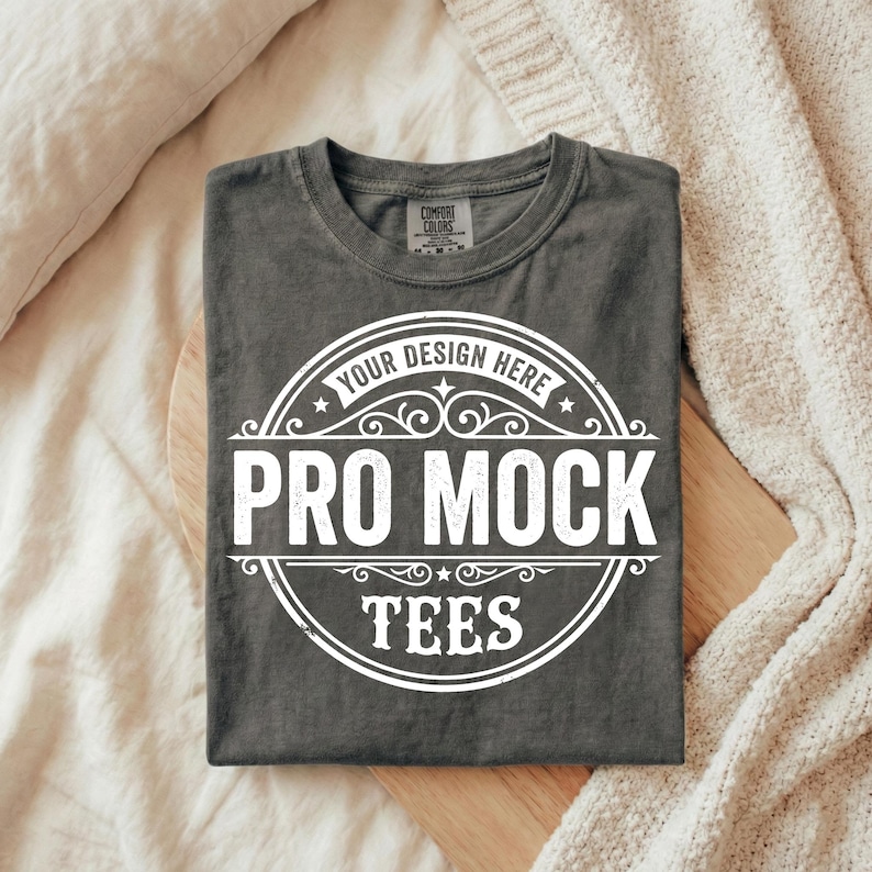 Puede incluir: Una camiseta gris carb&oacute;n doblada con un gr&aacute;fico circular blanco que dice "YOUR DESIGN HERE" en la parte superior y "PRO MOCK TEES" en letras may&uacute;sculas grandes. La camiseta est&aacute; sobre una superficie de madera, con una manta de color crema en el fondo.