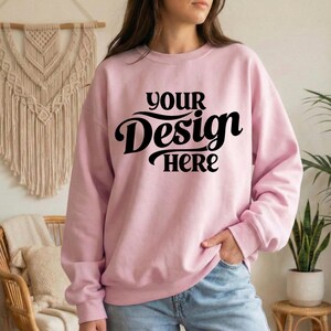 May include: A light pink sweatshirt with the text "YOUR Design HERE" in black script. The sweatshirt is a crew neck style and appears to be made of a soft material. The model is wearing blue jeans.
