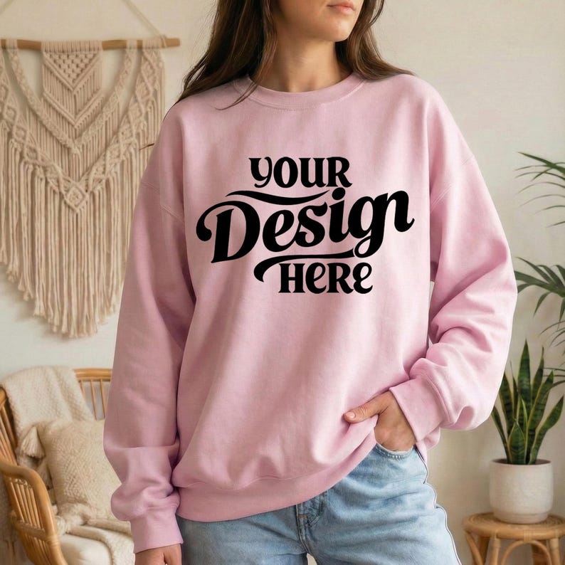 May include: A light pink sweatshirt with the text "YOUR Design HERE" in black script. The sweatshirt is a crew neck style and appears to be made of a soft material. The model is wearing blue jeans.