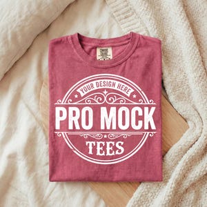 Puede incluir: Una camiseta burdeos doblada con un gr&aacute;fico circular blanco que dice "YOUR DESIGN HERE" y "PRO MOCK TEES". La camiseta est&aacute; sobre una superficie de madera, con una manta y una almohada beige en el fondo.