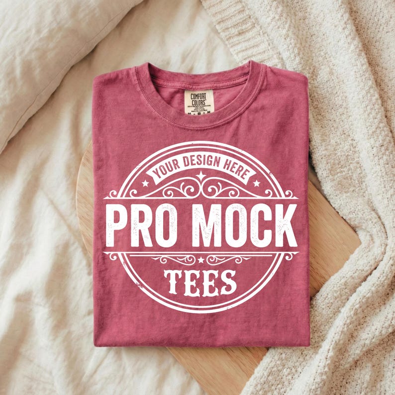 Puede incluir: Una camiseta burdeos doblada con un gr&aacute;fico circular blanco que dice "YOUR DESIGN HERE" y "PRO MOCK TEES". La camiseta est&aacute; sobre una superficie de madera, con una manta y una almohada beige en el fondo.