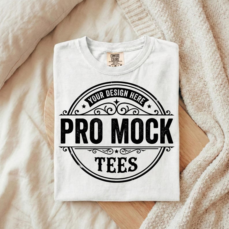 Puede incluir: Camiseta blanca con cuello redondo y dise&ntilde;o circular negro. El dise&ntilde;o incluye el texto "YOUR DESIGN HERE" en la parte superior y "PRO MOCK TEES" en letras negritas. La camiseta est&aacute; doblada sobre una superficie de madera.