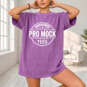 May include: A purple t-shirt with the text "PRO MOCK TEES" in a white circular design. The shirt is a solid color and has short sleeves. The model is wearing the shirt.