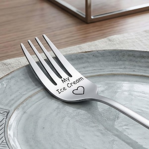 Puede incluir: Un tenedor de acero inoxidable con cuatro púas, apoyado sobre un plato azul claro. El tenedor tiene las palabras "My Ice Cream" y un símbolo de corazón grabado en el mango. El plato tiene un borde decorativo.