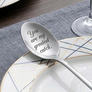 May include: A white ceramic mug with a black handle and the text "But First, Coffee" in a black, handwritten-style font. The mug is on a white surface.