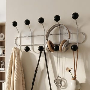 Puede incluir: Un perchero de pared con un marco horizontal de metal cromado y diez ganchos, cada uno rematado con una perilla esférica negra. El perchero sostiene unos auriculares beige, una correa de bolso negra, un collar con colgante plateado y un jarrón.