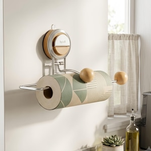 May include: A wall-mounted paper towel holder featuring a silver metal frame with a suction cup base labeled by'ell. The holder includes two light wood spheres at the ends of the horizontal bar, holding a roll of paper towels with a green geometric pattern.