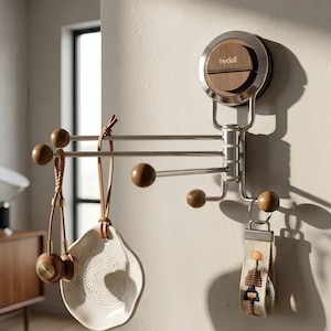 Puede incluir: Un perchero giratorio de pared con una base circular que muestra el texto "bydell" en blanco, un marco de metal plateado cepillado y varios brazos horizontales que terminan en esferas de madera de color marrón oscuro. Varios objetos cuelgan.