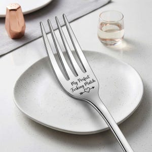 Puede incluir: Un tenedor de acero inoxidable pulido con cuatro púas, colocado sobre un plato blanco. El tenedor está grabado con la frase "My Perfect Forking Match" y un diseño de flecha-corazón. Un salero de cobre y un vaso pequeño con líquido están en el fondo.