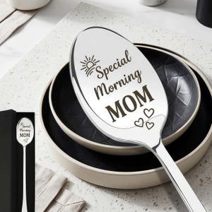 Puede incluir: Una cuchara plateada con las palabras "Special Morning MOM" grabadas, junto con un sol y corazones. La cuchara se muestra sobre un plato blanco y negro, con una caja de regalo negra en el fondo.