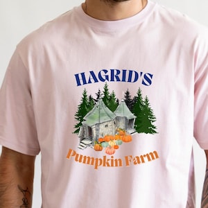 May include: A light pink t-shirt featuring the text "Hagrid's Pumpkin Farm" in blue and orange. The graphic includes a small house, pumpkins, and trees. The shirt is a crew neck and has short sleeves.