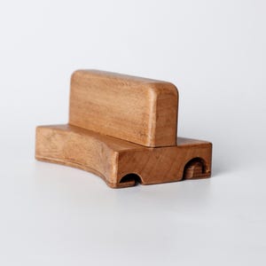 May include: A wooden toy car with a light brown finish. The toy has a rectangular body with rounded edges and a smaller rectangular block on top. The car has two circular cutouts on the front.