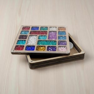 May include: A wooden bead organizer with a clear lid, filled with various colorful beads. The beads are arranged in rectangular compartments, showcasing a spectrum of colors including red, blue, green, and purple. The organizer sits atop a matching wooden base.