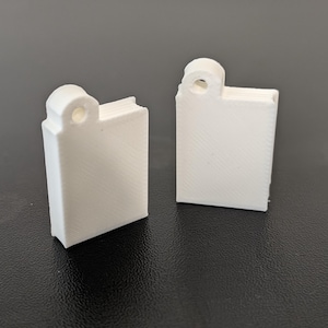 May include: Two white, rectangular, 3D-printed charms shaped like small books. Each charm features a vertical orientation with a rounded tab at the top containing a circular hole for attachment, and a textured surface showing fine diagonal print lines.