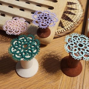 Könnte beinhalten: Nahaufnahme von handgefertigten Spitzendeckchen in Rosa, Lila, Grün und Hellblau, jeweils auf einer Holzspule befestigt. Die Deckchen weisen komplizierte Blumenmuster auf und zeigen die filigrane Handwerkskunst.