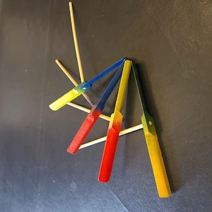 Könnte beinhalten: Eine Sammlung farbenfroher, handbemalter Cocktailspieße aus Holz. Die Spieße sind in Rot, Gelb, Blau und Grün bemalt und haben einen natürlichen Holzgriff. Die Spieße sind in einem Sternmuster vor einem dunklen Hintergrund angeordnet.