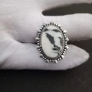 May include: Oval ring with a white stone and black markings, set in a silver metal frame. The ring has a decorative border with small, round accents. The stone's pattern resembles a natural landscape.