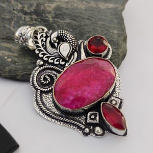 May include: A silver pendant with an oval, large, pink gemstone as the focal point. Two smaller red gemstones and ornate silver detailing surround the main stone. The pendant has a peacock design at the top.