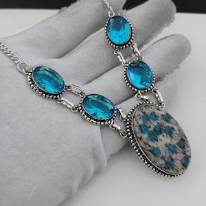 May include: A silver necklace with oval and round blue gemstones and a large oval pendant with a speckled blue and grey pattern. The necklace is displayed on a white surface.