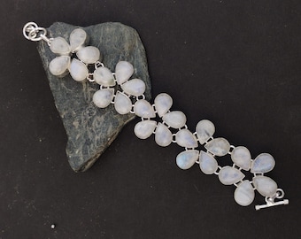 Op de afbeelding: Een zilveren armband met een bloemmotief, bezet met peervormige, iriserende maansteen edelstenen. De armband wordt getoond op een donkergrijze steen tegen een zwarte achtergrond. Het heeft een tuimelsluiting.