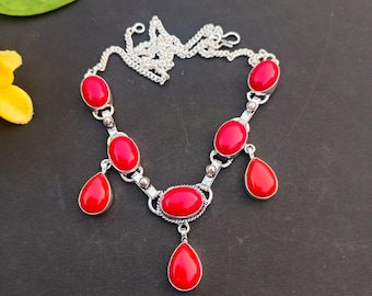 May include: A silver necklace featuring oval and teardrop-shaped red gemstones. The necklace has a chain and is set against a dark background. The gemstones are a vibrant red color, and the metal has a shiny finish.