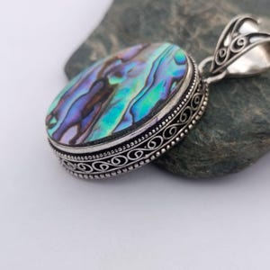 May include: Oval pendant with a vibrant abalone shell center, showcasing iridescent blues, greens, and purples. The shell is set in a silver-toned metal frame with an ornate, scrollwork design around the edge and bail. A unique piece of jewelry.