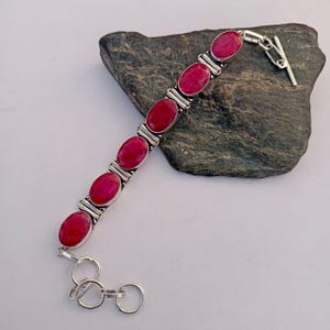 Op de afbeelding: Een zilveren armband met ovale, dieprode edelstenen. De armband heeft een tuimelsluiting en wordt gepresenteerd op een donkergrijze steen tegen een witte achtergrond. De edelstenen zijn gezet in zilvermetaal.