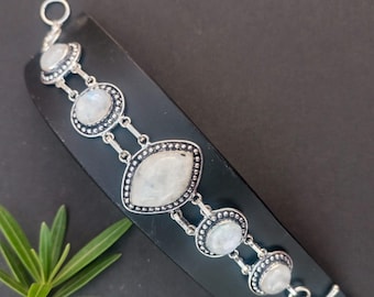 Op de afbeelding: Een zilverkleurige armband met maansteen cabochons. De armband heeft een ovale centrale steen en cirkelvormige stenen, allemaal gezet in een gedetailleerd, kralenomrand frame. De armband wordt op een zwart oppervlak gepresenteerd.