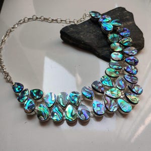 Puede incluir: Collar de plata con colgantes en forma de lágrima de concha de abulón iridiscente en tonos azules, verdes y morados. Los colgantes están engarzados en plata y unidos por una cadena de plata. El collar se exhibe sobre una piedra oscura.