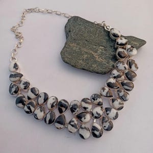 May include: A silver necklace with teardrop-shaped zebra jasper stones. The black and white stones are set in silver bezels and linked by a silver chain. The necklace is displayed on a white surface with a dark green rock in the background.