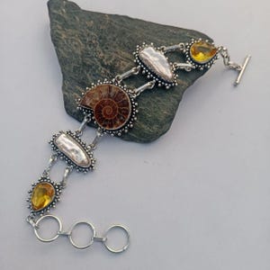May include: A silver bracelet featuring a fossilized ammonite, pear-shaped yellow gemstones, and white baroque pearls. The bracelet has a toggle clasp and is displayed on a gray stone.