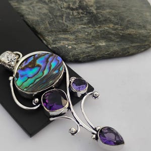 May include: A silver pendant featuring an oval abalone shell with iridescent blue and green hues, accented by three purple amethyst stones. The pendant has a decorative, flowing design.