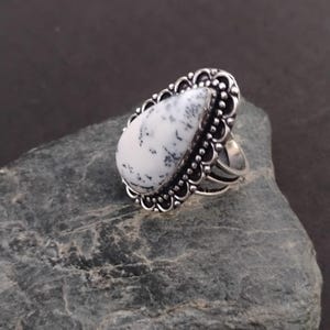 May include: A silver ring with a large, teardrop-shaped stone. The stone is white with black speckles and set in a decorative, scalloped silver frame. The ring sits on a grey, textured surface.