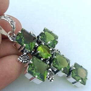 Op de afbeelding: Een zilverkleurige hanger in de vorm van een kruis, versierd met meerdere groene edelstenen. De edelstenen zijn een mix van vierkante en ovale slijpvormen, gezet in een gedetailleerd, sierlijk metalen ontwerp. De hanger heeft een lus voor een ketting.