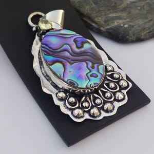 May include: A silver pendant featuring an oval abalone shell with shimmering blues, purples, and greens. The shell is set in a decorative silver frame with a small green gemstone. The pendant has an ornate design with detailed silverwork.