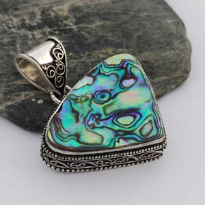 May include: Abalone shell pendant with a silver bail. The iridescent shell displays swirls of green, blue, and purple. The pendant has a triangular shape with a decorative silver border.