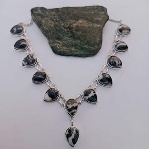 May include: A silver necklace with teardrop-shaped black and white patterned stones. The stones are set in a silver metal frame and connected by silver chain links. The necklace has a central pendant with a larger teardrop stone.