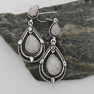 May include: Silver-coloured earrings with a teardrop design. Each earring has a large, pear-shaped, off-white stone, set within a decorative silver frame. The earrings have a hook closure and are shown on a dark grey stone surface.