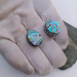 Puede incluir: Un par de pendientes de plata con concha de abulón iridiscente en un engaste redondo. La concha muestra tonos de azul, verde y morado. Los pendientes tienen un borde decorativo y un cierre de gancho simple.